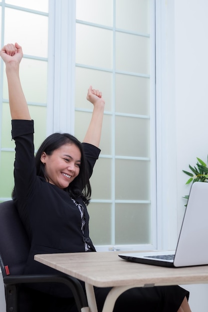 Business woman sitting on chair raising both hands with success expression in front of laptop