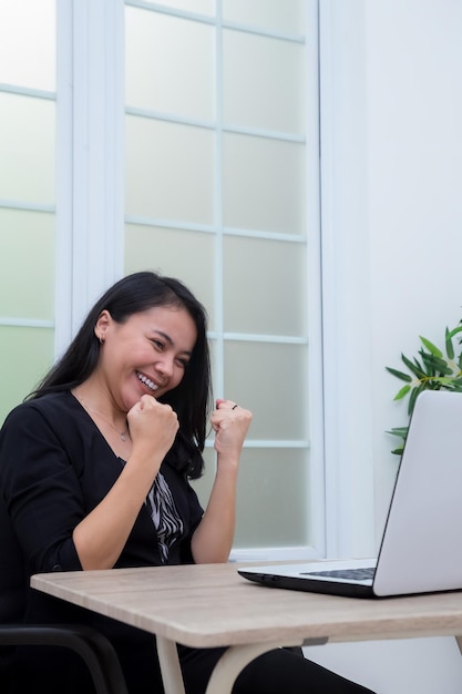 Business woman sitting on chair while raising both hands with success expression in front of laptop