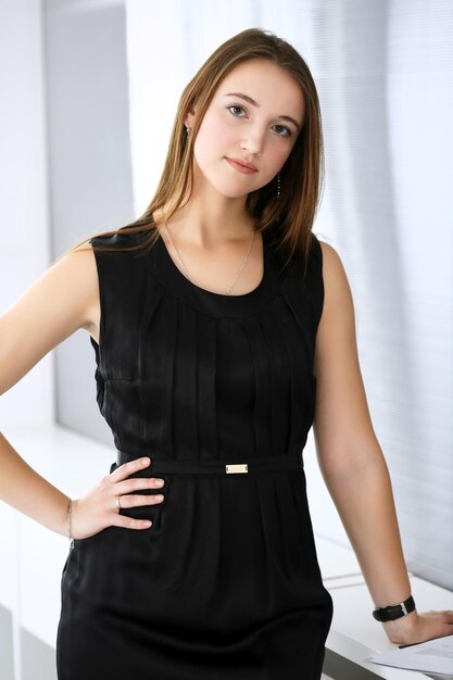 Business woman standing and looking at camera in office of highlight background Headshot of beautiful girl in black dress