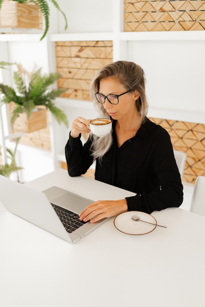 Business woman in stylish casual outfit sitting in office and using lap top and drinking coffe