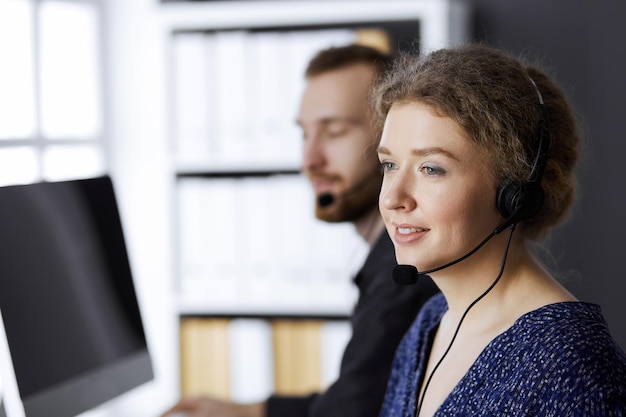 Business woman talking by headset while sitting with red-bearded colleague in modern office. Telemarketing and customer service. Diverse people group in call center.