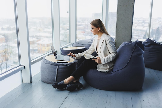 Business woman thinks while writing in a notebook at work Modern successful woman Young smiling girl in glasses sits in a chair at a table in an open office