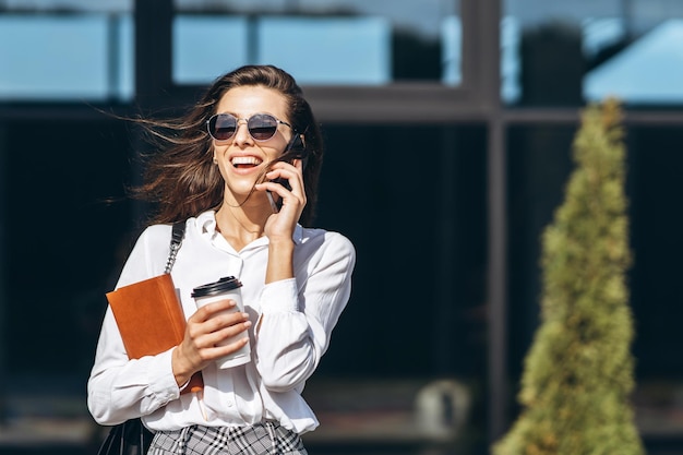 Business woman walking and talking on the cellphone outdoors near modern business center with notebook