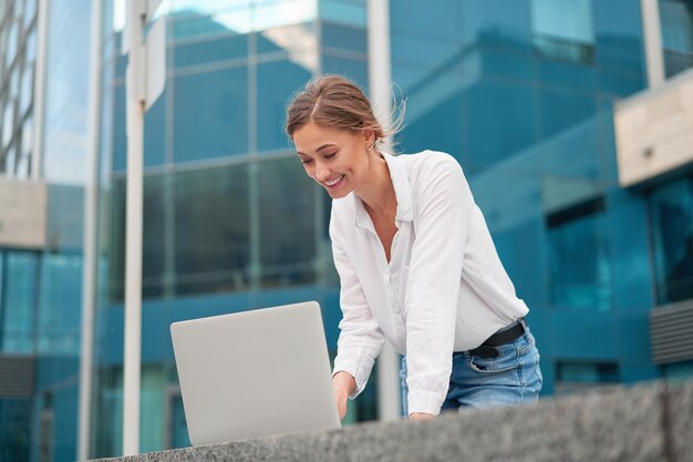 Business woman in a white blouse uses a laptop to work while outside, working outdoors. In the background is a business center with large windows.