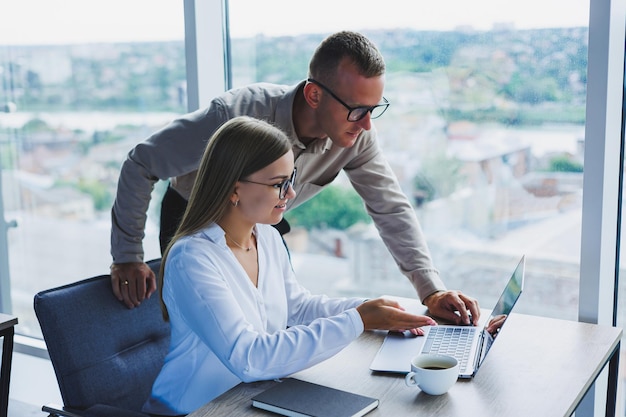 Business woman with a laptop for a male colleague who looks into an open notebook Business cooperation and teamwork European millennials at the table in the office Modern successful people