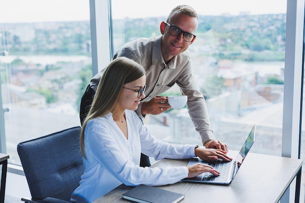 Business woman with a laptop for a male colleague who looks into an open notebook Business cooperation and teamwork European millennials at the table in the office Modern successful people