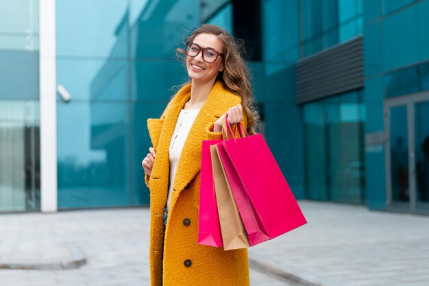 Business woman with shopping bags dressed yellow coat walking outdoors corporative building background Caucasian female business person near office building Stylish businesswoman season sale