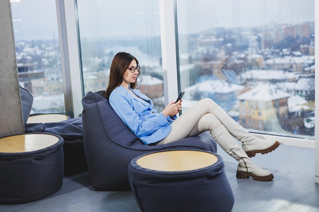 Business young woman in glasses with long dark hair in casual clothes smiling and looking at the phone browsing the smartphone during a day off in the workspace