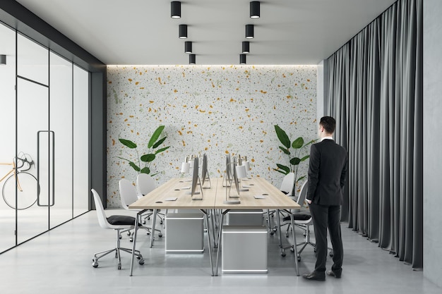 Businessman back view in modern board room with light wooden conference table surrounded by chairs green plants near multi colored wall and dark curtain