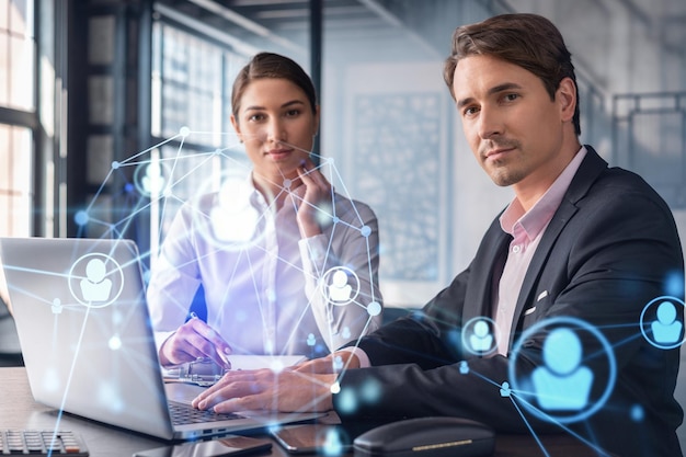 Businessman and businesswoman in formal wear working together looking for new employees to hire for international business consulting HR social media icons over modern panoramic office background