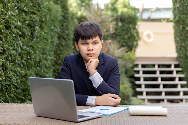 Businessman concept a young businessman in navy suit sitting in the garden with the documents and the laptop on the table looking thoughtful and serious.