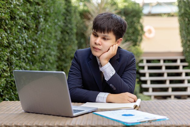 Businessman concept a young businessman in navy suit sitting in the garden with the documents and the laptop on the table looking thoughtful and serious.