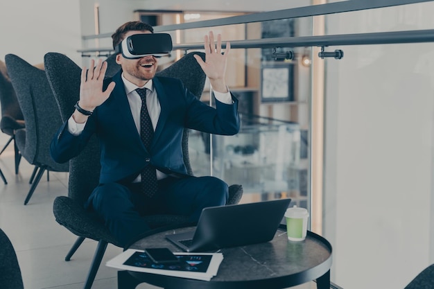 Businessman dressed in formal suit wearing VR goggles while working on laptop in cafe