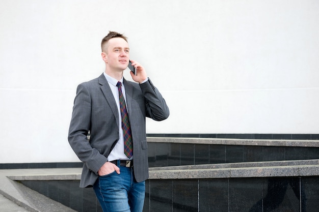 Businessman, dressed in grey jacket, blue jeans, white shirt and tie comes out of buisness center and talking on the phone while walking