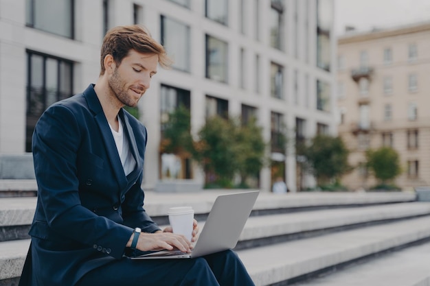 Businessman in formal suit reads news via laptop computer works online drinks takeaway coffee