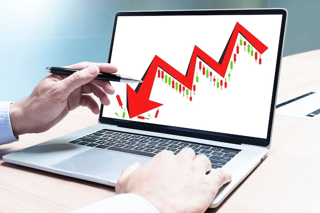 Businessman hand holding pen to display laptop with red arrow downtrend stock chart on display