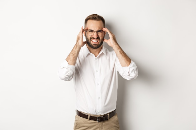 Businessman having headache, grimacing and holding hands on head, standing over white background.