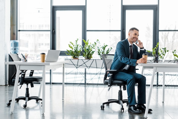 businessman holding paper cup while talking on smartphone in modern office