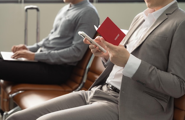 Businessman holding passport sitting at airport terminal and checking for flight on mobile phone