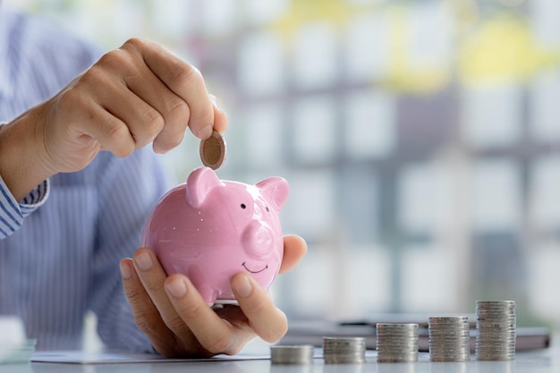 A businessman is putting coins into a pink pig piggy bank he is saving for investments money growth stock investments The idea of investing money to make it worthwhile to grow and be sustainable