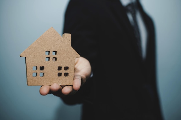 Businessman in modern black suit hand giving house model to customer on dark background, financial business, buy house, real estate broker, finance, home loan contract, buy and sell house concept