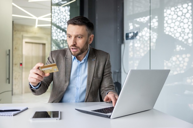 Businessman in modern office appears shocked while holding credit card and viewing laptop indicating