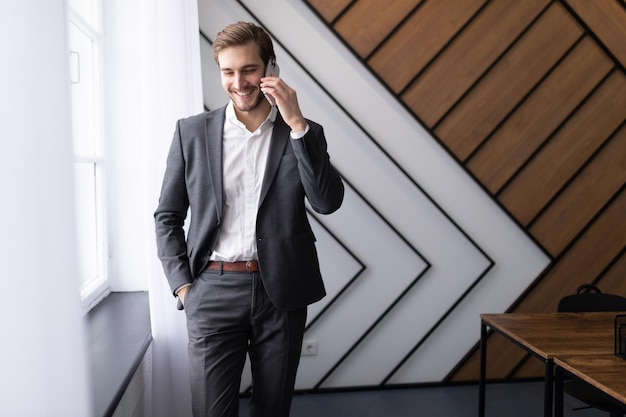 A businessman in an office at the window with a smile on his face speaks on a mobile phone in a