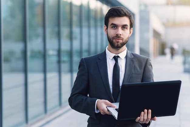 Businessman outdoors on a background of cityscapes holding a laptop looking ahead. 