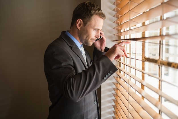 Businessman peeking through blinds while on call