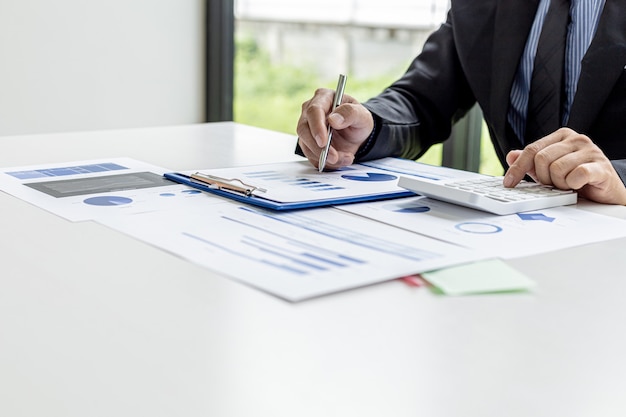 Businessman presses a white calculator, he is the owner of the company, he is checking company financial documents in his office, financial documents show chart format. Concept of financial management