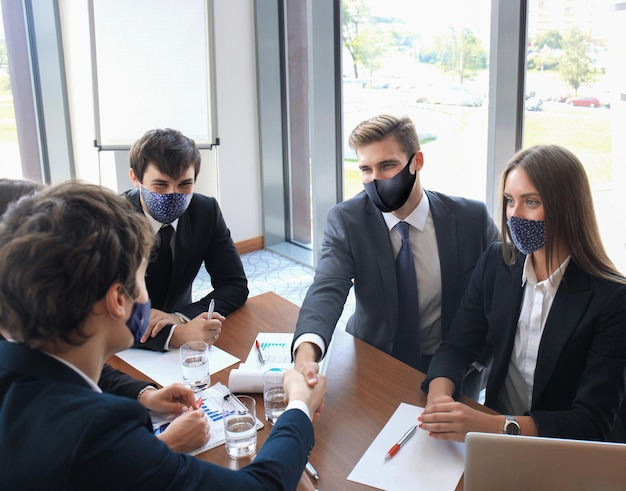 Businessman in preventive mask shaking hands to seal a deal with his partner and colleagues who also wear preventive masks in office.