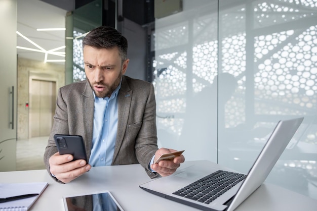 Businessman shocked by fraud notification while holding credit card and phone in office setting with