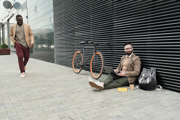 Businessman sitting on the ground and working on laptop with other man walking along the street