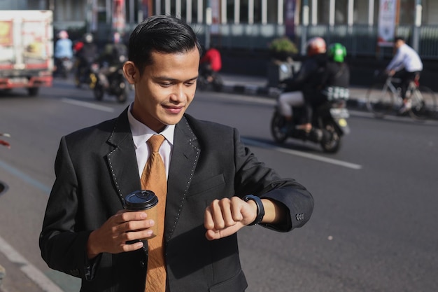 Businessman standing in the city street with coffee cup and checking the time on watch