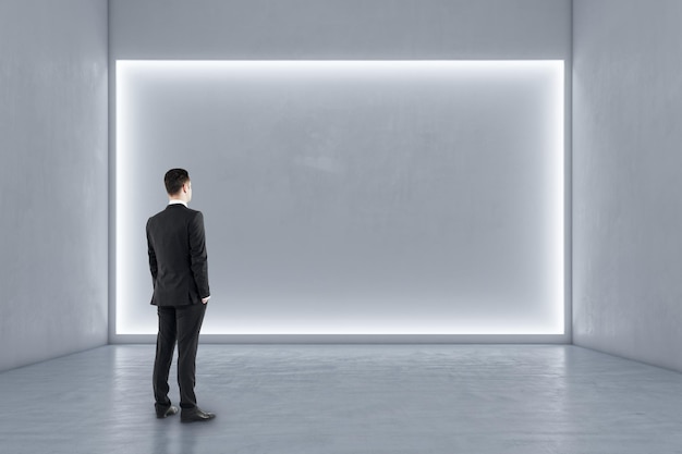 Businessman standing in modern exhibition hall interior with illuminated white mock up wall and concrete floor Gallery concept