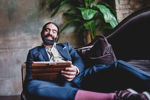 Businessman in suit relaxing on the couch in workspace using a tablet smiling and happily reading