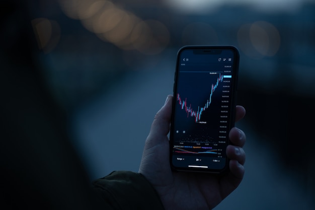 Businessman or trader using mobile phone to check real time stock market data while standing outdoors, selective focus on male hand holding smartphone with forex graph chart