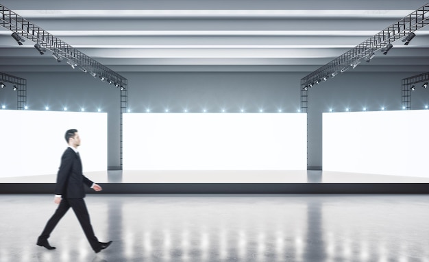 Photo businessman waiks in empty light showroom with blank white glowing boards and black stage in the center