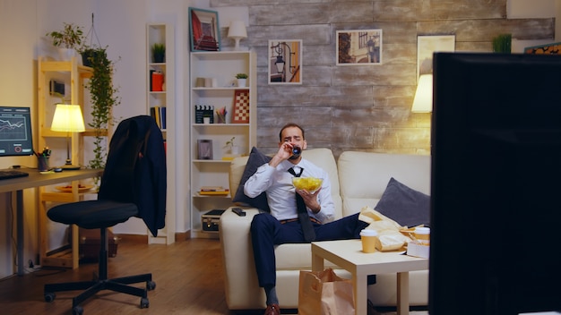 Businessman with tie relaxing on couch after a long day at work eating chips and watching tv.