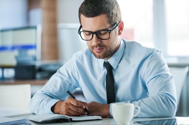 Businessman at work. Confident young businessman in formalwear writing in note pad while sitting at his working place