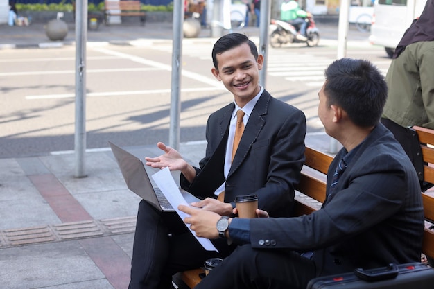 businessman working on laptop and his partner showing paperwork while sitting together outside