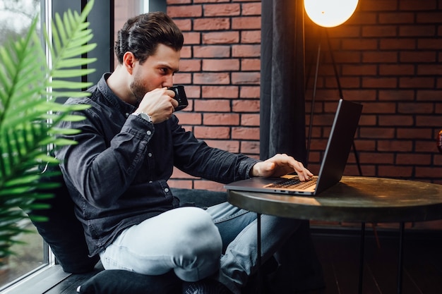 Businessman working on laptop, holding cup with coffee or latte in modern cafe near windows.