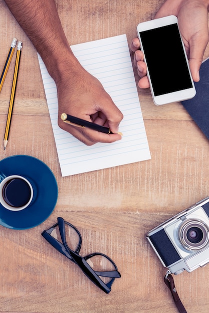 Businessman writing on notepad while holding smart phone by camera and eyeglasses on table