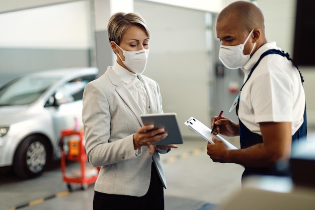 Businesswoman and African American mechanic wearing face masks and cooperating while using touchpad in a workshop