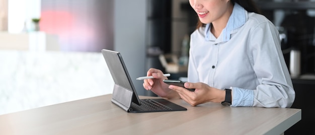 Businesswoman holding mobile phone and searching information on computer tablet at office desk.