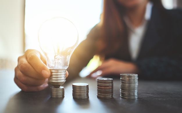 Businesswoman holding and putting lightbulb on coins stack on table for saving energy and money concept