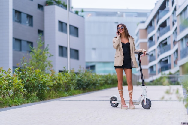 A businesswoman moving with an electric scooter around the city sunglasses smiling talking on the phone