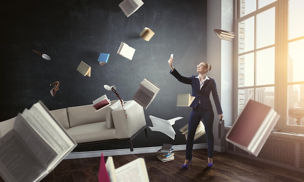 Businesswoman standing and making selfie with flying books all around her. Mixed media