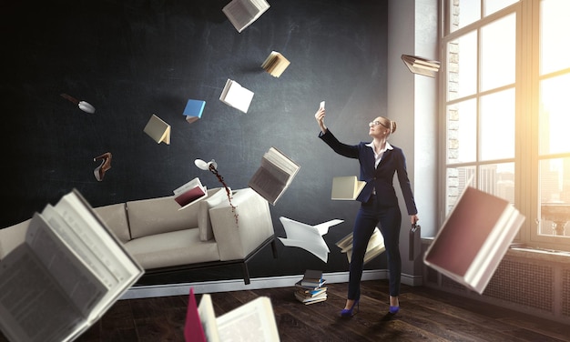 Businesswoman standing and making selfie with flying books all around her. Mixed media