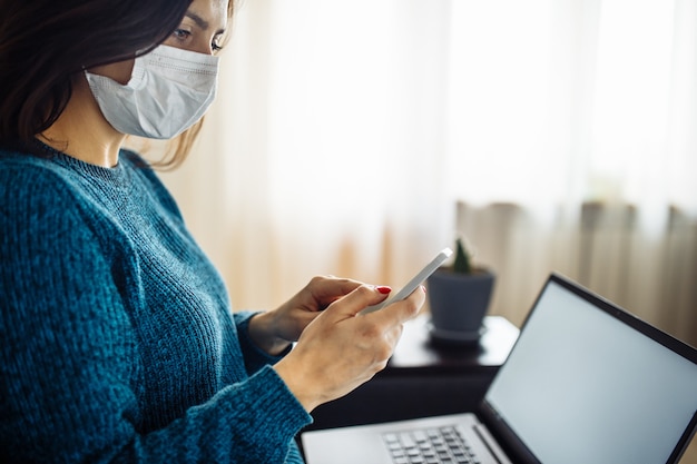 Businesswoman stays home and works during coronavirus epidemia quarantine. Female worker wearing a medical mask and holding her cell phone in the hands. Covid-19 pandemia spread prevention concept.